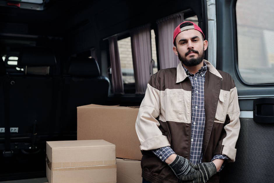 A young man with a beard, wearing a red baseball cap backward, a multicolored jacket, a plaid shirt, and gloves, stands inside the open rear of a van during a home relocation. The van's interior is li