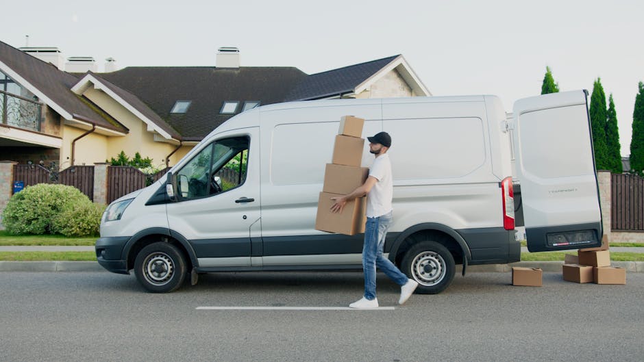 A young man with a beard, wearing a red baseball cap backward, a multicolored jacket, a plaid shirt, and gloves, stands inside the open rear of a van during a home relocation. The van's interior is li