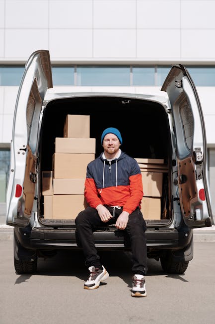A man with a beard, wearing a blue beanie, a navy hoodie with red and gray sections, and black pants, is sitting on the edge of an open vehicle trunk at a loading area. The trunk is filled with several medium-sized cardboard boxes arranged in stacks, some placed directly on the vehicle floor and others on top of each other. The vehicle is a light-colored van, positioned on a paved surface in front of a modern building with a white tiled exterior. The interior of the van is visible through the open rear doors, revealing additional boxes and some packing materials. The lighting is natural, highlighting the details of the boxes, the man's clothing, and the vehicle. This scene depicts the process of home relocation or furniture transport as part of a professional removals service, with Man With a Van East Ham providing inside and outside loading support.