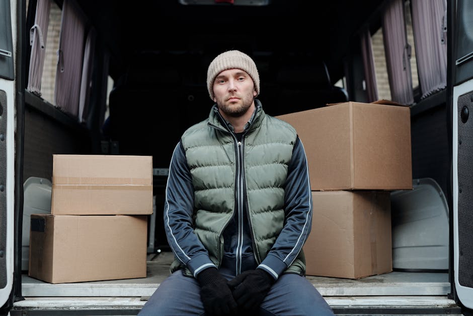 A man with a beard, wearing a blue beanie, a navy hoodie with red and gray sections, and black pants, is sitting on the edge of an open vehicle trunk at a loading area. The trunk is filled with several medium-sized cardboard boxes arranged in stacks, some placed directly on the vehicle floor and others on top of each other. The vehicle is a light-colored van, positioned on a paved surface in front of a modern building with a white tiled exterior. The interior of the van is visible through the open rear doors, revealing additional boxes and some packing materials. The lighting is natural, highlighting the details of the boxes, the man's clothing, and the vehicle. This scene depicts the process of home relocation or furniture transport as part of a professional removals service, with Man With a Van East Ham providing inside and outside loading support.