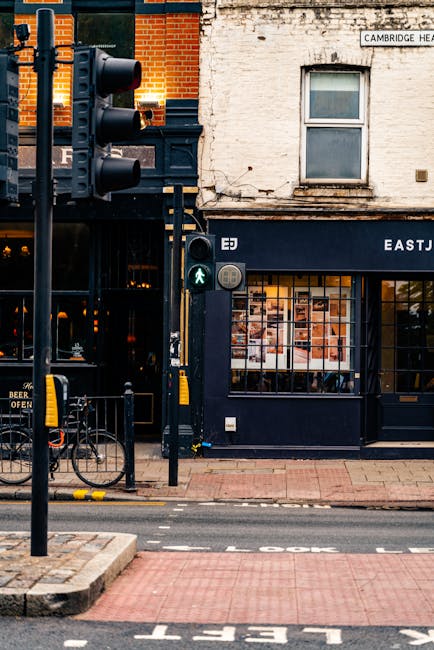 A traffic light with a green pedestrian signal is positioned on a sidewalk next to a black standing sign and a bicycle rack, all in front of a building with a white brick facade and large glass windows. The building features a dark blue shopfront with a sign that partially reads 'EAST J...' and behind the windows, there are images or posters visible inside. The surrounding environment includes a paved pavement and a road with a red tactile paving strip and white markings. This scene is captured during daylight, with natural light illuminating the area. The image may be associated with house removals or moving services, as it depicts an urban street scene relevant to relocation logistics, although no people or moving activity are visible in this specific shot.