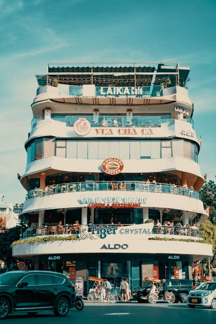 A multi-storey building with a distinctive curved design, featuring several floors with glass railings and outdoor terraces. The ground floor houses retail shops, including an ALDO store, while the upper levels appear to be commercial or hospitality spaces, with visible signage for cafes and restaurants such as Laika Cafe and a burger restaurant. The building's exterior combines white and pastel colours, with some greenery on the terraces. In the foreground, there are parked cars, motorcycles, and pedestrians on the street, indicating a busy urban environment typical of East Ham. Natural daylight and clear weather illuminate the scene, highlighting the building's architectural details and the activity along the pavement, which may include furniture and boxes associated with a home or business move. Man With a Van East Ham may be involved in such relocations, supporting efficient furniture transport and packing during house removals or commercial relocations.