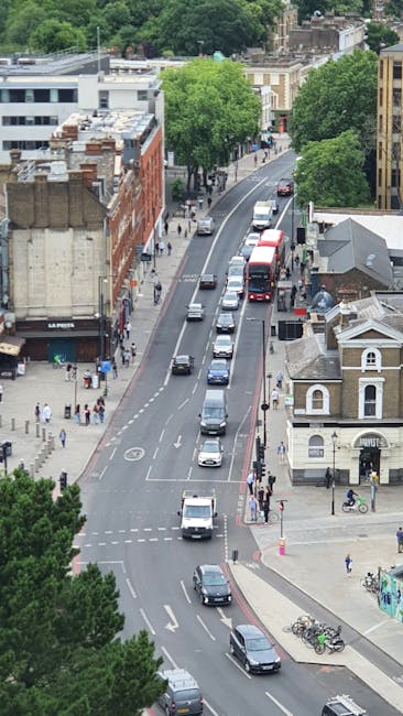 A view from an elevated perspective of a busy street in East Ham, showing multiple vehicles including cars and a red double-decker bus moving along the road. On the left side, a sidewalk is populated with pedestrians, some walking and others waiting at the crossing. To the right, a small building with decorative architecture and a bicycle rack with several bikes are visible near the corner. Large, leafy green trees line the street, providing shade and separation between the road and surrounding buildings, which include a mix of older brick structures and modern office blocks. The area exhibits typical urban activity associated with home relocation and furniture transport logistics, as part of a house removal process, with the scene capturing the bustling environment typical of East Ham during a packing and moving operation.