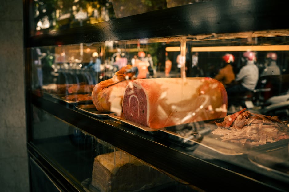 A large cut of raw beef, wrapped partially in plastic and resting on a metal tray inside a glass display fridge, with visible marbling and a deep red interior. The reflection on the glass shows an outdoor scene with blurred figures of people and motorcycles, suggesting a busy street environment. This image is part of a house removal context, where packing or preparing fresh ingredients might be managed during relocation. The setting indicates a focus on food storage or catering, relevant to moving services that handle perishable goods. The fridge’s interior lighting highlights the texture and colour of the meat, and the scene implies careful handling during the packing process for a house move. Man With a Van East Ham occasionally manages these details as part of their home relocation and furniture transport services, ensuring all household items, including food items, are properly prepared for a seamless move.