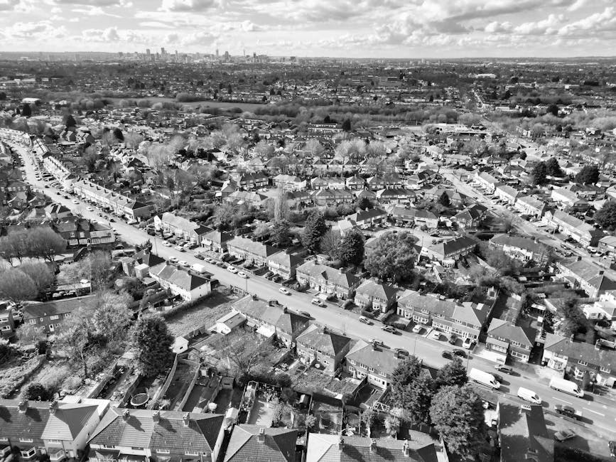 A view from an elevated perspective of a busy street in East Ham, showing multiple vehicles including cars and a red double-decker bus moving along the road. On the left side, a sidewalk is populated with pedestrians, some walking and others waiting at the crossing. To the right, a small building with decorative architecture and a bicycle rack with several bikes are visible near the corner. Large, leafy green trees line the street, providing shade and separation between the road and surrounding buildings, which include a mix of older brick structures and modern office blocks. The area exhibits typical urban activity associated with home relocation and furniture transport logistics, as part of a house removal process, with the scene capturing the bustling environment typical of East Ham during a packing and moving operation.