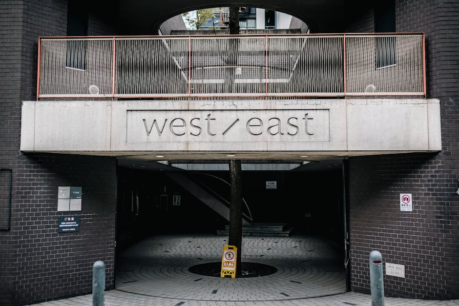 The image shows the exterior of a multi-story residential building with a concrete balcony featuring a metal railing painted in a muted pink color, positioned above a central driveway entrance. The building's facade is made of dark brown brick, contrasting with the light grey concrete of the balcony. The balcony has a decorative panel with vertical metal bars and small square windows visible on either side. Beneath the balcony, there is an opening leading into an underground parking or stairwell area, which is dark and partially obscured. Close to the entrance, there are signage boards, one of which is blue, indicating parking restrictions or building information, and another showing a no-smoking symbol. A yellow and black safety sign is placed on a tree or pole directly in front of the opening, while bollards are positioned on either side of the driveway to prevent vehicle intrusion. The scene is set during daytime with natural lighting, illustrating an urban environment suitable for home relocation or furniture transport activities associated with house removals by Man With a Van East Ham.