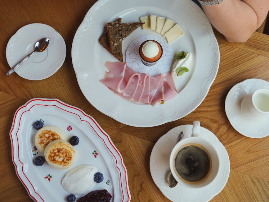 A top-down view of a wooden table set with breakfast items and dessert plates. In the upper center, a white ceramic plate displays sliced cheese, a boiled egg in an egg cup, slices of ham, a piece of bread, and some leafy garnishes. To the left, a small white saucer holds a polished silver spoon resting on a paper napkin. Below, an oval-shaped white plate features two small pancakes topped with caramelized sugar, accompanied by fresh blueberries, a dollop of cream, and a serving of berry jam. To the right, a white ceramic cup filled with black coffee is placed on a matching saucer with a small teaspoon, and beside it is a white jug. The table surface has a natural wood finish, and part of a person's arm is visible at the top right. This setting reflects a breakfast scene, captured clearly for accessibility and supporting themes of home life.