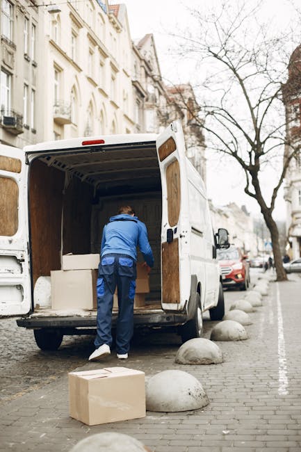 A man wearing a blue jacket and dark trousers is seen loading large cardboard boxes and other packing materials into the back of a white cargo van parked on a city street. The van's rear doors are open, revealing the interior with wooden panels, and the scene suggests a home relocation process involving furniture transport and packing. The street is lined with parked cars, including a red vehicle, and buildings with multiple stories, typical of an urban environment, are visible in the background. The cobblestone pavement features rounded concrete bollards separating the parking area from the sidewalk, and a leafless tree is seen in the background. This scene illustrates the logistics of packing and loading for house removals, with tools like blankets or straps possibly used for secure transport, supporting professional removals services such as those offered by Man With a Van East Ham.