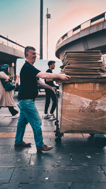 A man from Man With a Van East Ham is engaged in a home relocation process, standing outdoors on a paved area under an overpass during daytime. He is pushing a large, metal trolley loaded with multiple stacked cardboard boxes and large flat-packed items wrapped in transparent plastic film, indicating packing and moving activities. The trolley is positioned near the edge of a moving van or transport vehicle, partially visible in the background, used for furniture transport. Other pedestrians are visible in the background, walking past the scene. The environment includes concrete paving, metal railings on the overpass, and a lamppost, all contributing to the urban setting typical of east London. The photograph captures the loading process involved in house removals, highlighting furniture and boxes being prepared for transportation during a scheduled move, matching the context of professional removals services offered by [COMPANY_NAME].