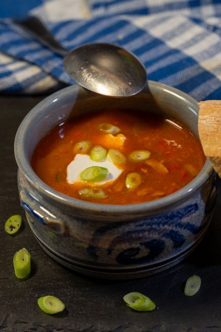 A ceramic bowl filled with tomato-based soup containing chopped green onions and a dollop of white cream or sour cream, placed on a dark textured surface. A metal spoon rests on the edge of the bowl, with some green onion slices scattered around the bowl. The bowl is situated inside a home, reminiscent of a kitchen or dining area, with a striped blue and white cloth partially visible in the background. This scene captures the preparation or presentation of a warm, homemade meal, aligning with home-related activities such as packing or moving, as it showcases everyday kitchenware and ingredients. The natural lighting highlights the rich colour and texture of the soup, emphasizing a homely and inviting atmosphere, suitable for visually supporting content about house moves or kitchen packing in East Ham.