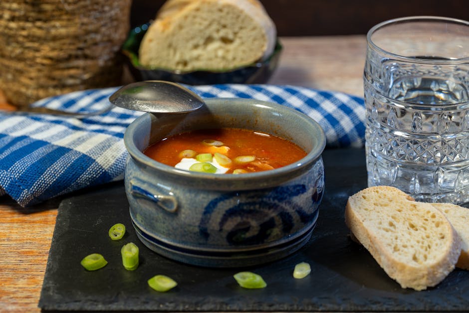 A close-up view of a rustic table setting featuring a bowl of tomato soup garnished with chopped green onions, placed on a dark slate serving board. To the right of the soup, there is a transparent, textured glass of water, partially filled. Behind the soup, a partially sliced piece of white bread with a soft interior and crusted exterior rests on the table, along with an open bread roll in the background. A folded blue and white checkered cloth napkin is beneath the bread, with a wooden surface visible underneath. The scene suggests a casual, home-cooked meal with natural lighting, and the setting could be part of a relocation or moving process where packing and meal preparation are involved, aligning with services offered by Man With a Van East Ham during house removals and furniture transport.