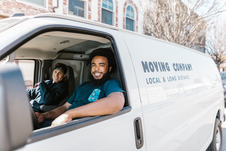 Inside the cab of a white moving van from Man With a Van East Ham, two men are seated, preparing for a home relocation. One man with a beard and dark curly hair is smiling at the camera, wearing a dark blue t-shirt, while the other man, with lighter hair and a headband, is seated behind him in the driver’s seat, also smiling. The van’s interior shows various packing materials, including cardboard boxes, plastic wrapping, and furniture blankets, indicating ongoing packing and loading activities. The exterior of the van has the company's logo and the text 'MOVING COMPANY LOCAL & LONG DISTANCE' printed on the side. The van is parked outside a residential area with multi-storey buildings, and the scene suggests an active loading process typical of furniture transport and house removals, supported by the company's professional services.