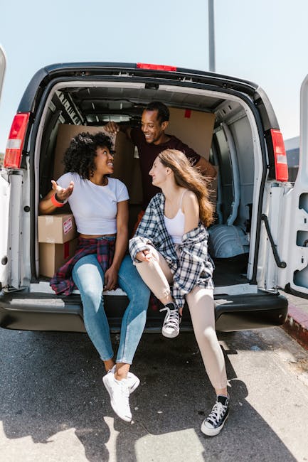 Three people, two women and one man, sit inside the open rear of a grey moving van, engaged in a conversation and smiling. The woman on the left has curly black hair, is wearing a white T-shirt, jeans, and white sneakers, with her hands gesturing as she talks. The woman on the right has long brown hair, wearing a checked shirt tied around her waist, a white tank top, and black sneakers, with one knee raised. The man stands inside the van, leaning slightly forward with a smile, wearing a dark shirt. Visible inside the van are cardboard boxes, a wrapped parcel, and packing materials, indicating a home relocation. The setting appears to be outdoors on a driveway or street, with part of the van's interior and the pavement visible. The scene captures a moment of packing, loading, or a break during the furniture and box loading process, which aligns with house removals and moving services provided by Man With a Van East Ham.