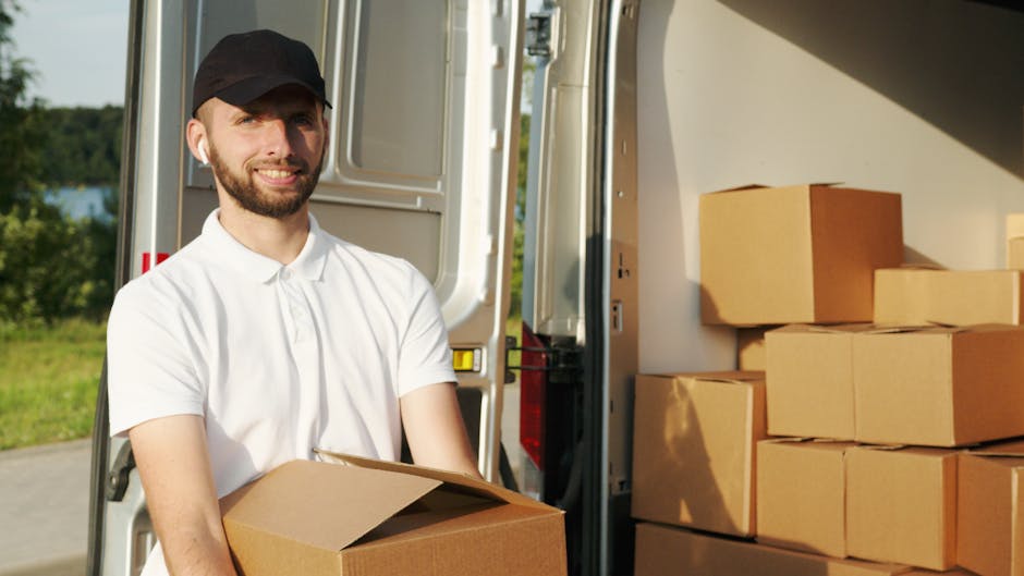 Inside the cab of a white moving van from Man With a Van East Ham, two men are seated, preparing for a home relocation. One man with a beard and dark curly hair is smiling at the camera, wearing a dark blue t-shirt, while the other man, with lighter hair and a headband, is seated behind him in the driver’s seat, also smiling. The van’s interior shows various packing materials, including cardboard boxes, plastic wrapping, and furniture blankets, indicating ongoing packing and loading activities. The exterior of the van has the company's logo and the text 'MOVING COMPANY LOCAL & LONG DISTANCE' printed on the side. The van is parked outside a residential area with multi-storey buildings, and the scene suggests an active loading process typical of furniture transport and house removals, supported by the company's professional services.