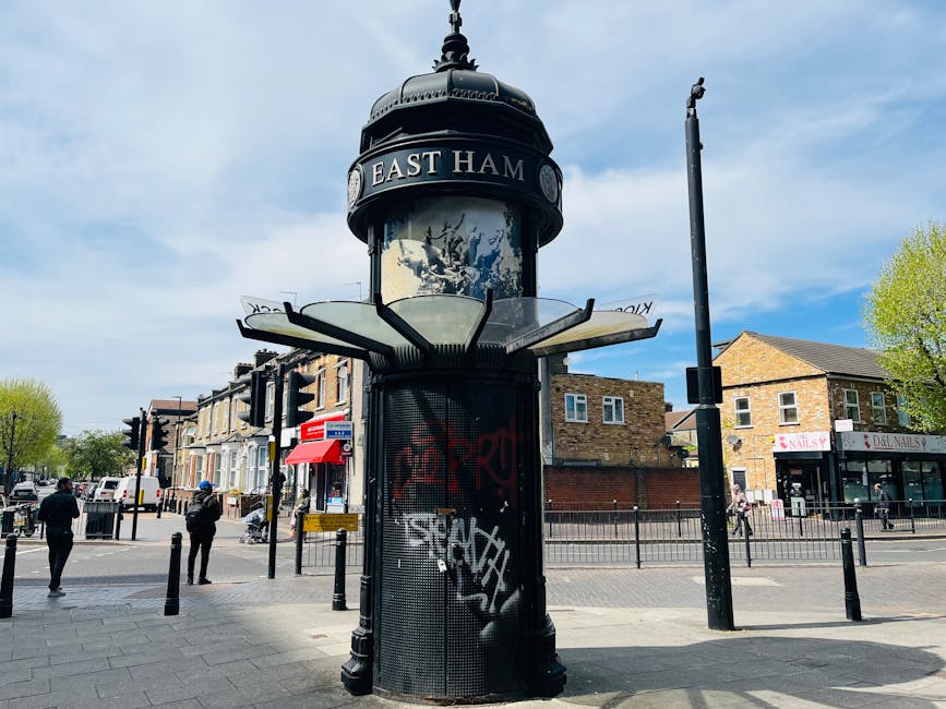 A street scene in East Ham featuring a vintage-style, cylindrical pedestrian kiosk with a domed, black metal roof and a decorative finial on top. The kiosk displays the name 'EAST HAM' on a circular sign above the glass-panelled upper section, which appears to be partially obscured by reflective surfaces. Surrounding the kiosk, there are several black metal bollards along the pavement to prevent vehicle access. In the background, there are row of shops including a nail salon and a deli, with pedestrians walking nearby on a partly cloudy day, with some individuals standing and others walking along the street. The scene includes a tall, vertical pole with a street sign or lamp attached, and the overall environment is typical of a busy urban shopping area. This setting is relevant for discussions on urban removal challenges, such as navigating tight spaces or access restrictions during home relocation or furniture transport services in East Ham, as provided by Man With a Van East Ham.