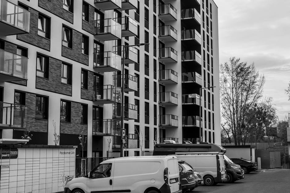 A black-and-white photograph of a modern multi-storey residential building with multiple balconies, each featuring glass railings and some with outdoor furniture or plants. Several cars, including a white van, a small utility vehicle, and other parked vehicles are positioned in the foreground on a paved area near the building. The vehicles are situated close to the building's entrance and loading zone, with some doors open as if in the process of loading or unloading. The building's exterior showcases a mix of brickwork and smooth wall panels, with tall, leafless trees visible on the right side, indicating seasonally sparse foliage. The lighting suggests daytime with a cloudy sky overhead. This scene depicts the logistical challenges of house removals and furniture transport in an urban environment, with [COMPANY_NAME] providing professional moving services within such settings, including access to upper floors and safety considerations during the loading process.