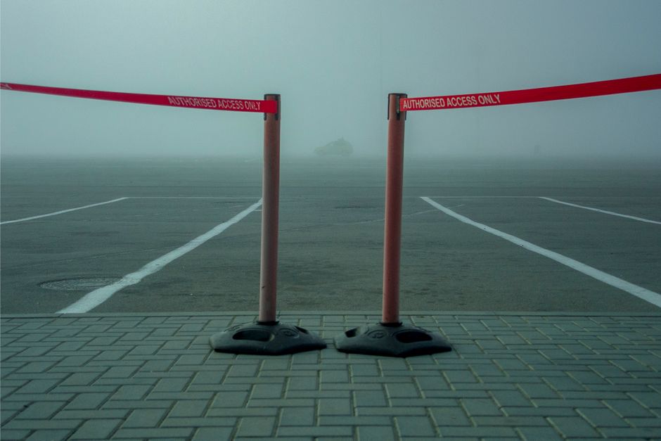 A foggy outdoor scene shows empty parking spaces with white lines on a paved surface, partially obscured by dense mist. In the foreground, two orange plastic barriers with black bases are positioned on a tiled sidewalk, connected by red tape marked with 'Authorised Access Only.' Behind the barriers, the parking area extends into the fog, with no visible vehicles or objects except for a faint, distant shape of a small boat or structure barely visible through the haze. The scene suggests restricted vehicle access due to parking suspensions, relevant in the context of house removals and furniture transport, with the barriers indicating a controlled loading bay area as might be seen during a home relocation or moving process managed by Man With a Van East Ham.
