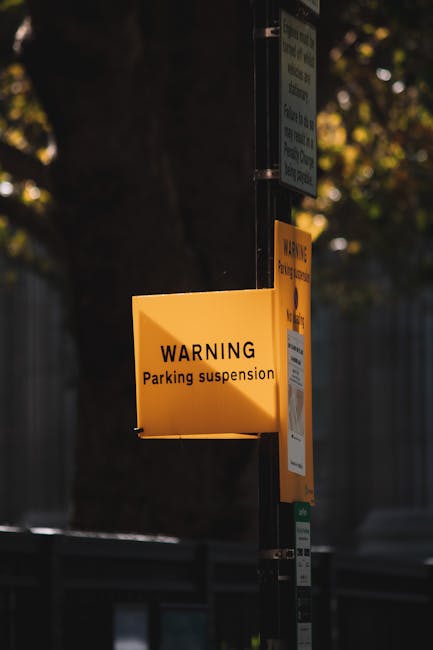 A foggy outdoor scene shows empty parking spaces with white lines on a paved surface, partially obscured by dense mist. In the foreground, two orange plastic barriers with black bases are positioned on a tiled sidewalk, connected by red tape marked with 'Authorised Access Only.' Behind the barriers, the parking area extends into the fog, with no visible vehicles or objects except for a faint, distant shape of a small boat or structure barely visible through the haze. The scene suggests restricted vehicle access due to parking suspensions, relevant in the context of house removals and furniture transport, with the barriers indicating a controlled loading bay area as might be seen during a home relocation or moving process managed by Man With a Van East Ham.