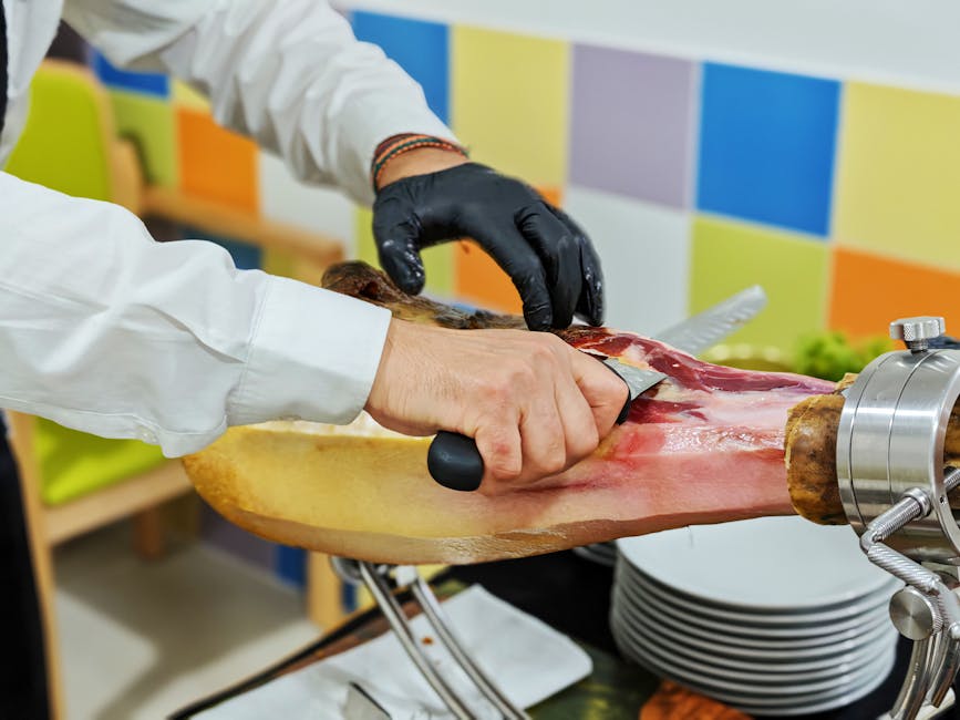 A person wearing black gloves and a white coat is slicing a large, cured leg of ham mounted on a professional meat carving stand, using a small knife. The carving process takes place indoors, in front of a brightly colored, checkered wall composed of yellow, blue, orange, and purple squares. The ham is placed on a wooden base, and there are stacked white plates beside it, with some serving as a catch tray for the sliced meat. This scene appears to be part of food preparation, but the equipment and setting bear resemblance to those used in commercial kitchens or specialty delicatessens, similar to how items might be packed and prepared for transport or display. The presence of a carving stand and utensils highlights precise slicing involved in high-quality meat service, aligning with professional handling during home relocation or catering events by companies like Man With a Van East Ham, which offers removals and related services.