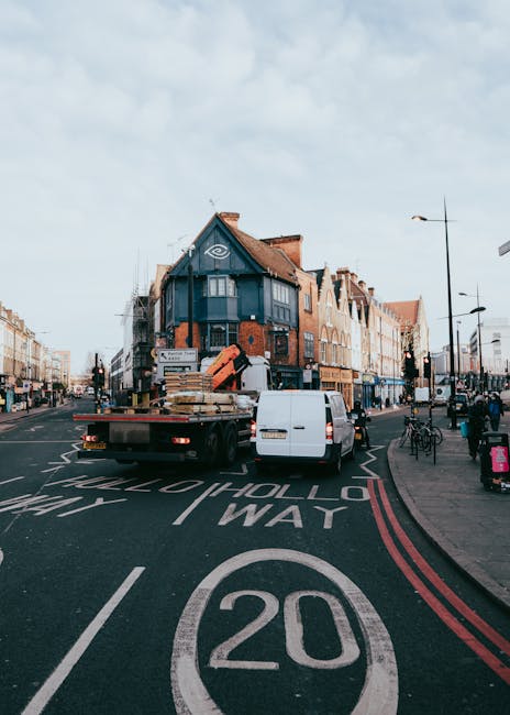 A white flatbed truck with wooden pallets loaded on its bed is parked on a one-way street in East Ham, London, during daylight hours. The pallets contain cardboard boxes and other packing materials, some wrapped in plastic film. The truck is in front of a building with a blue and brick facade, situated at a street corner with shops and apartments visible. A white van is parked behind the truck, and bicycles are secured to bike racks along the pavement. Street lamps, traffic signs, and road markings, including a 20 mph speed limit and a lane designation, are visible in the foreground, indicating an organized urban environment suitable for house removals and home relocation services. The scene suggests an active loading process in preparation for furniture transport, with clear weather providing good visibility for the ongoing moving operation, possibly facilitated by [COMPANY_NAME] for efficient packing and transportation.
