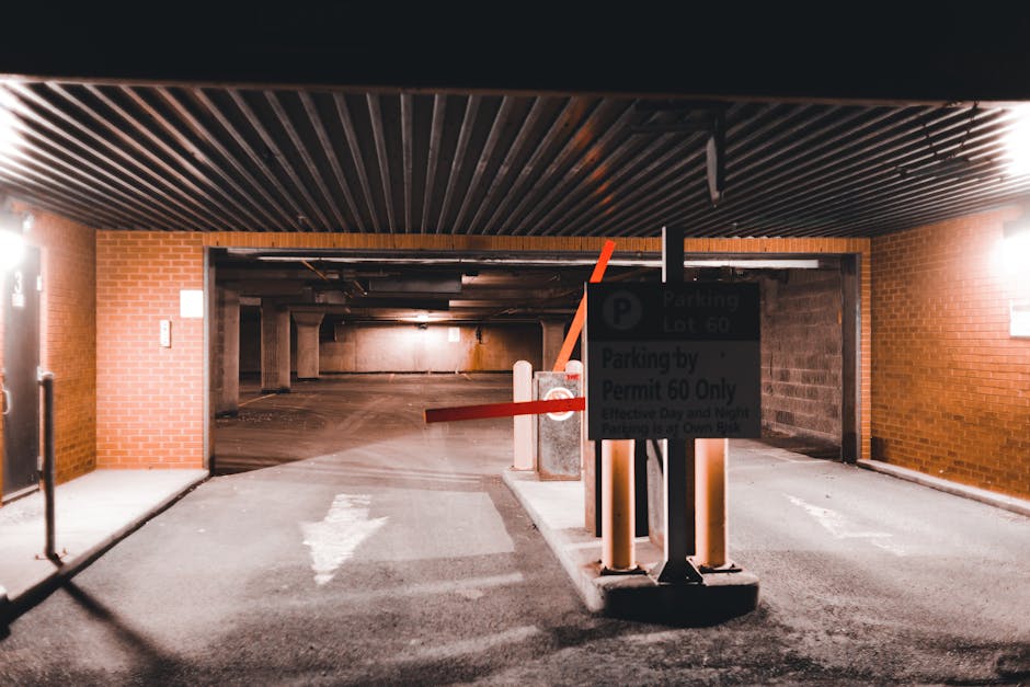 The image shows the entrance to an underground parking garage with a black ceiling and brick walls on either side. A gate with a red barrier arm is positioned across the driveway, which is marked with a white arrow pointing forward. There is a parking regulation sign mounted on a post in the center of the entrance area, indicating parking restrictions by permit only. Inside the garage, visible in the background, are concrete pillars and a dimly lit space indicating an accessible parking area. The surrounding environment appears clean and organized, consistent with a managed parking facility used for house removals or furniture transport. This setting reflects the type of parking considerations relevant for home relocation and professional removals services, such as those provided by Man With a Van East Ham. The image captures the process of parking lot access, essential for efficient loading and unloading during house moving or packing and moving procedures.
