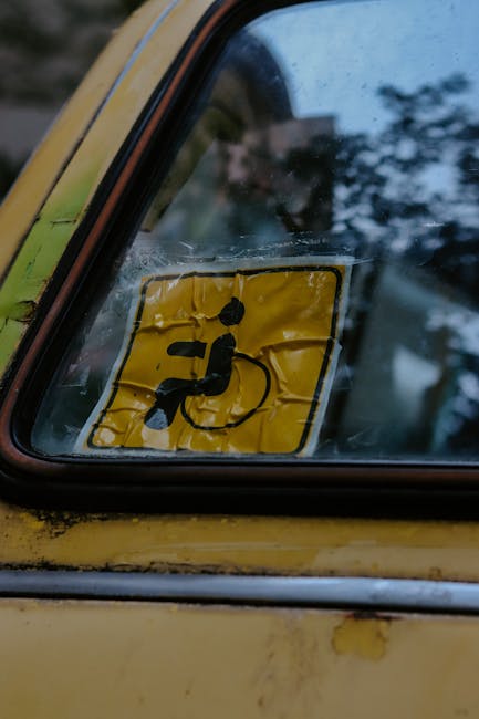 Close-up image of a yellow vehicle window with visible rain droplets, featuring a black and yellow disability parking permit sticker affixed to the glass. The sticker displays an icon of a person in a wheelchair and is partially wrinkled. The vehicle's exterior shows signs of weathering, with chipped paint and rust spots near the window frame. The background reflects an outdoor environment with blurred greenery and sky, indicating overcast weather. This image captures the context of transportation and vehicle access, relevant to house removal and furniture transport services as part of a home relocation process, such as those offered by Man With a Van East Ham.