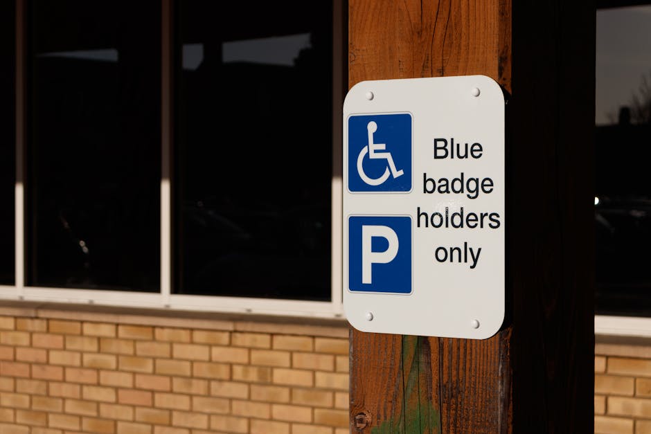 The image shows the entrance to an underground parking garage with a black ceiling and brick walls on either side. A gate with a red barrier arm is positioned across the driveway, which is marked with a white arrow pointing forward. There is a parking regulation sign mounted on a post in the center of the entrance area, indicating parking restrictions by permit only. Inside the garage, visible in the background, are concrete pillars and a dimly lit space indicating an accessible parking area. The surrounding environment appears clean and organized, consistent with a managed parking facility used for house removals or furniture transport. This setting reflects the type of parking considerations relevant for home relocation and professional removals services, such as those provided by Man With a Van East Ham. The image captures the process of parking lot access, essential for efficient loading and unloading during house moving or packing and moving procedures.